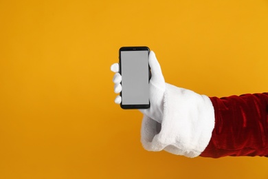 Photo of Santa holding modern mobile phone with blank screen on orange background, closeup. Space for text