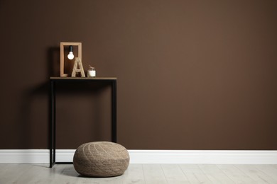 Comfortable knitted pouf and table with decor elements near brown wall indoors, space for text. Interior design