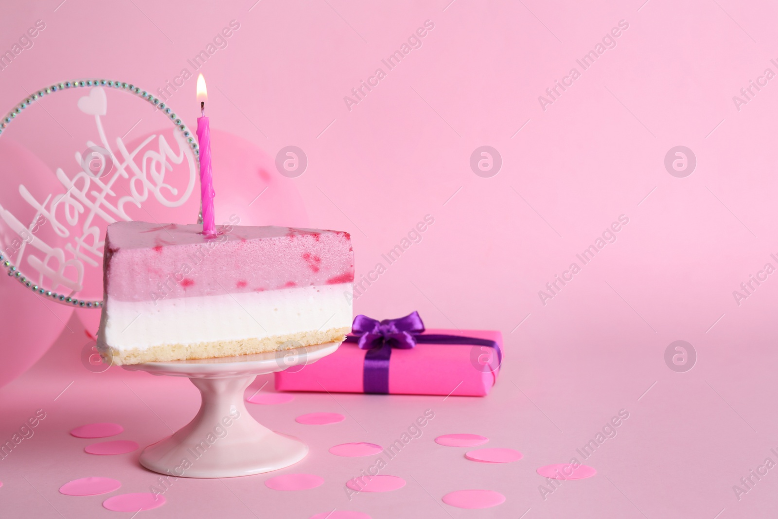 Photo of Piece of delicious birthday cake with candle near gift box and balloons on pink background, space for text