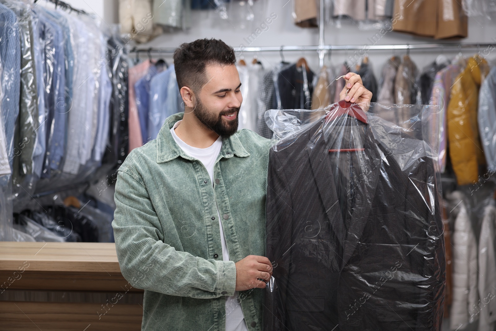 Photo of Dry-cleaning service. Happy man holding hanger with jacket in plastic bag indoors