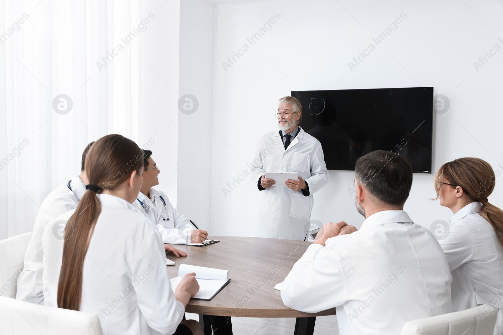 Photo of Team of doctors listening to senior speaker report near tv screen in meeting room. Medical conference