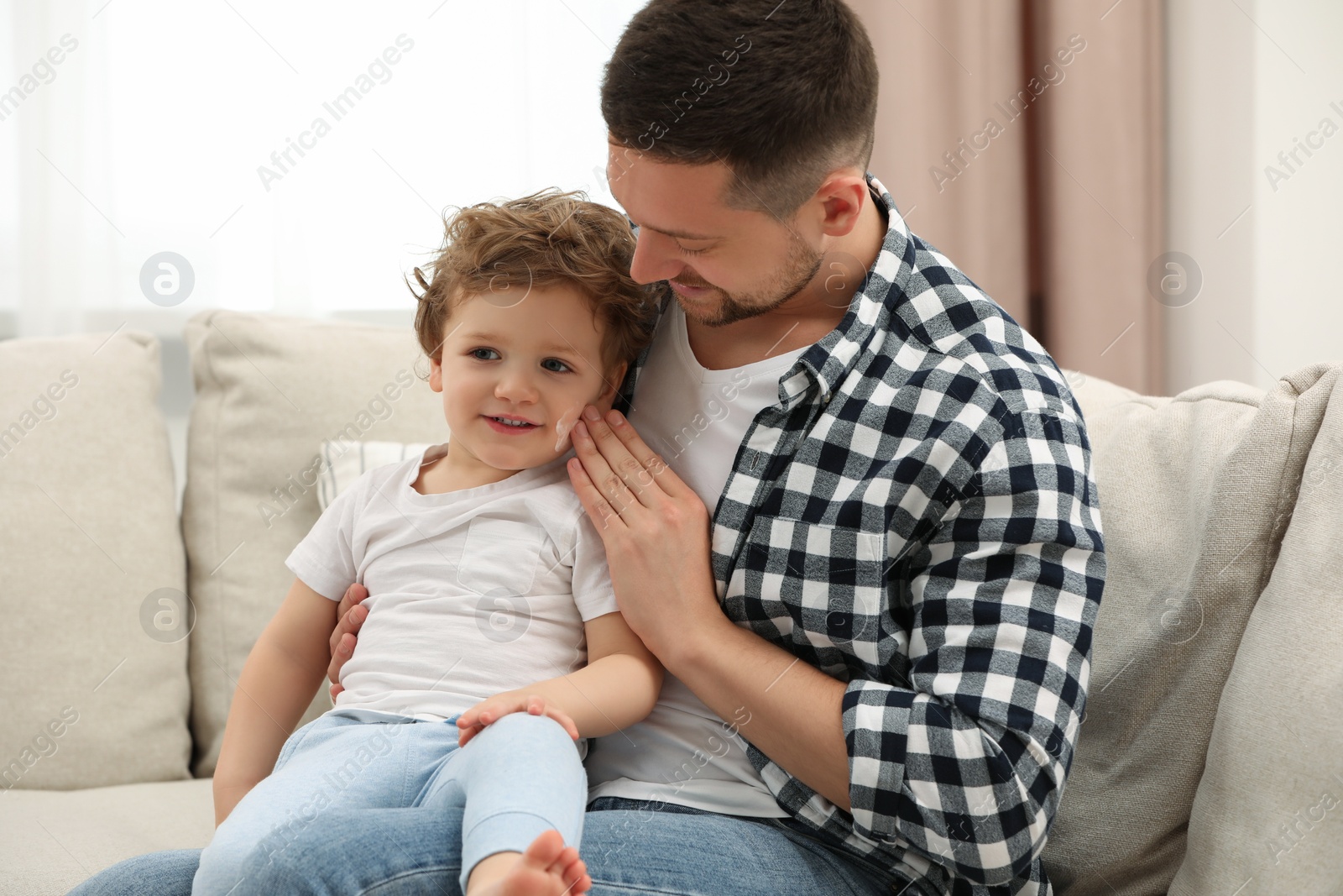 Photo of Father applying ointment onto his son`s cheek on sofa at home