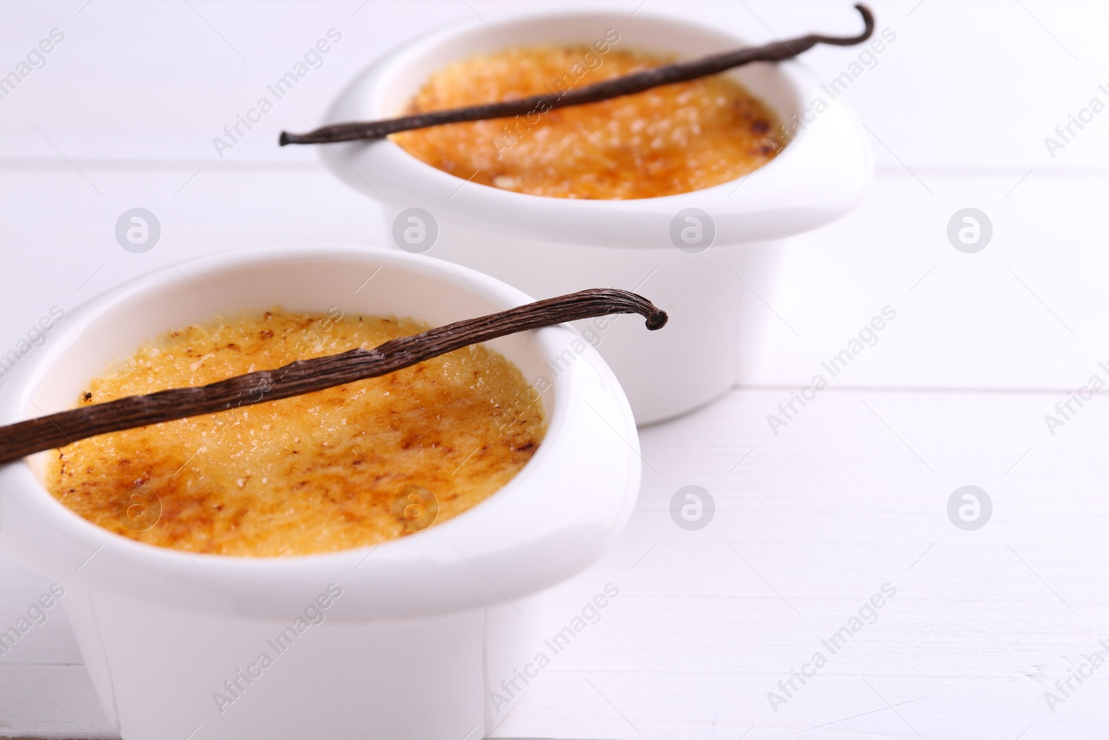 Photo of Delicious creme brulee in bowls and vanilla pods on white wooden table, closeup