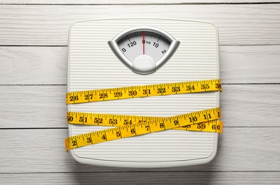 Photo of Weigh scales tied with measuring tape on white wooden table, top view. Overweight concept