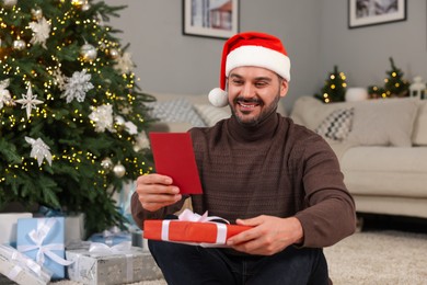 Photo of Happy man in Santa hat with Christmas gift reading greeting card at home