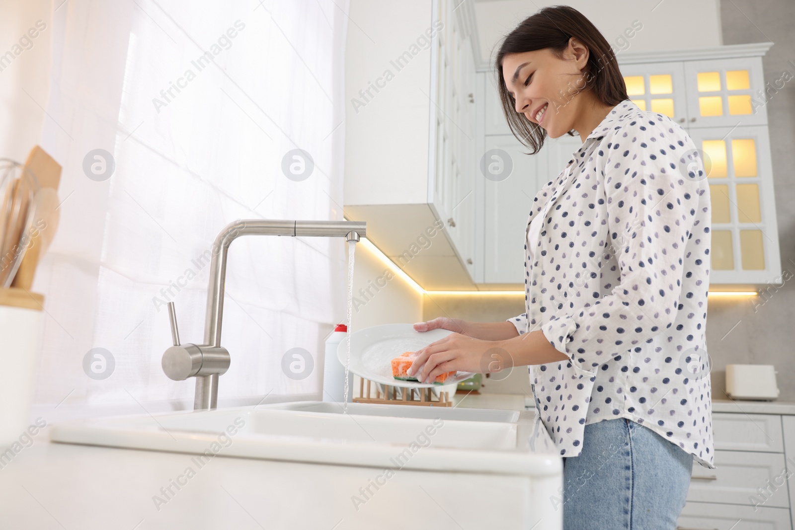 Photo of Happy young woman washing plate above sink in modern kitchen