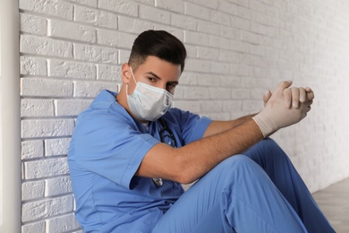 Sad doctor sitting near white brick wall indoors. Stress of health care workers during coronavirus pandemic