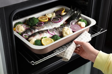 Photo of Woman putting baking tray with sea bass fish and vegetables into oven, closeup