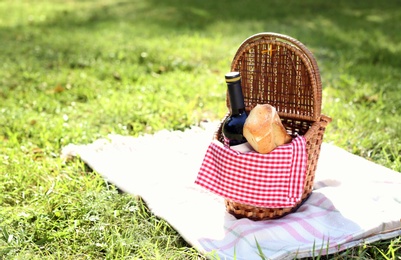 Photo of Wicker picnic basket with bottle of wine and bread on blanket in park. Space for text