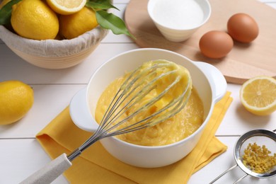 Photo of Delicious lemon curd in bowl, ingredients, whisk and sieve on white wooden table, closeup