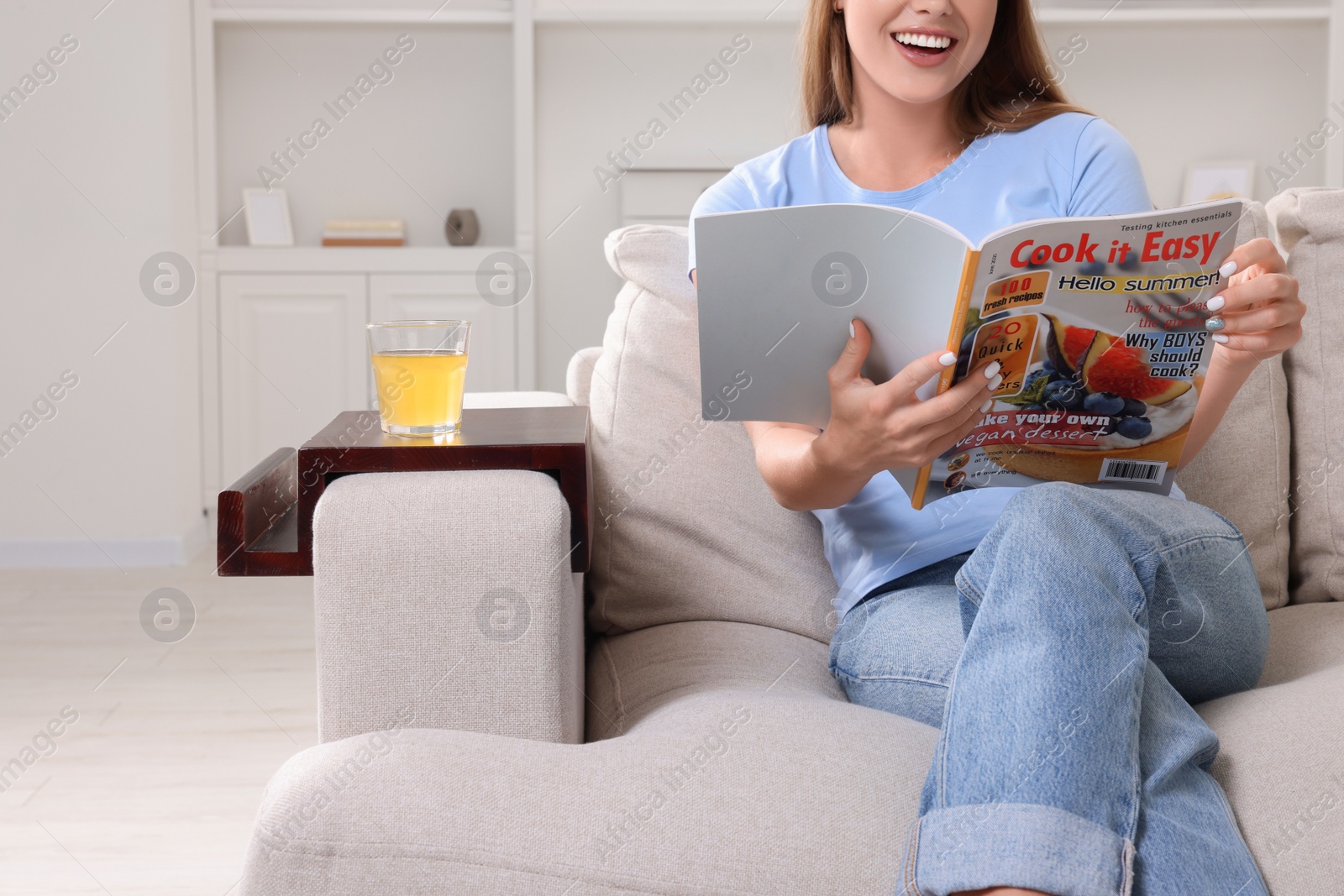 Photo of Glass of juice on wooden sofa armrest table. Woman reading magazine at home, closeup