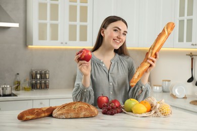 Woman with baguette, apple and string bag of fresh fruits at light marble table in kitchen