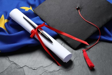 Photo of Graduation cap, diploma, pen and flag of European Union on black table