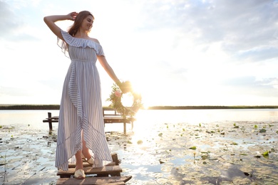 Photo of Young woman holding wreath made of beautiful flowers on pier near river