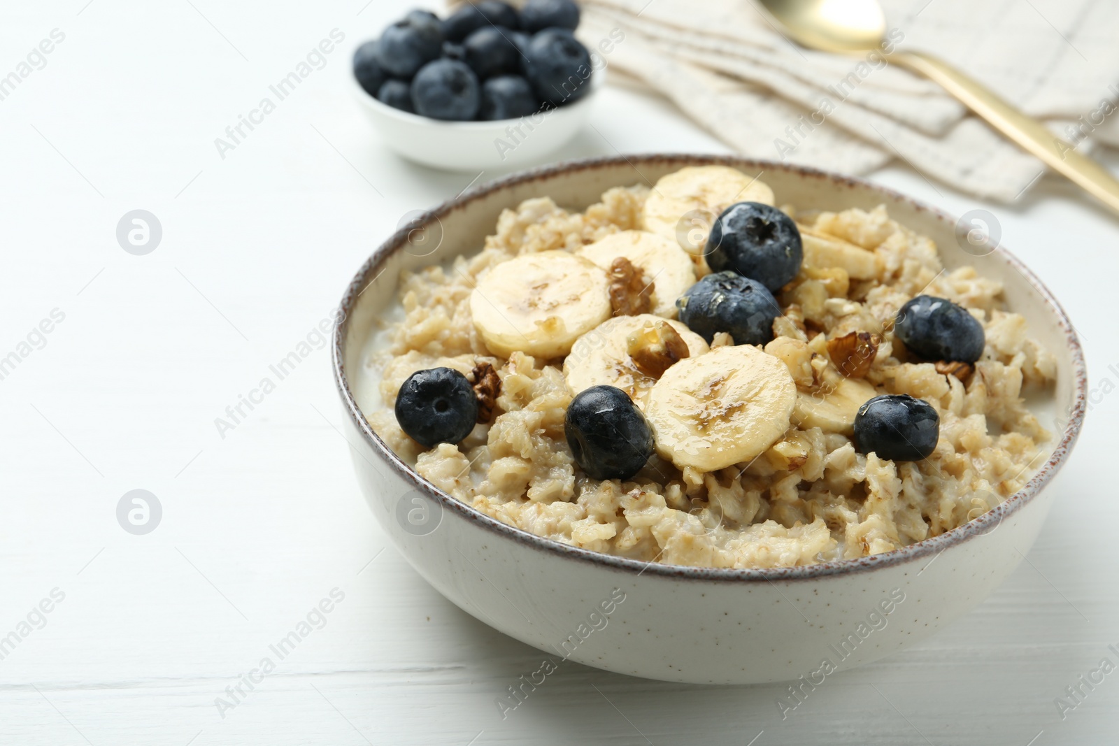 Photo of Tasty oatmeal with banana, blueberries, walnuts and honey served in bowl on white wooden table, space for text