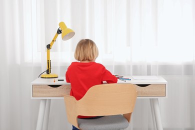 Photo of Little boy sitting at desk in room, back view. Home workplace
