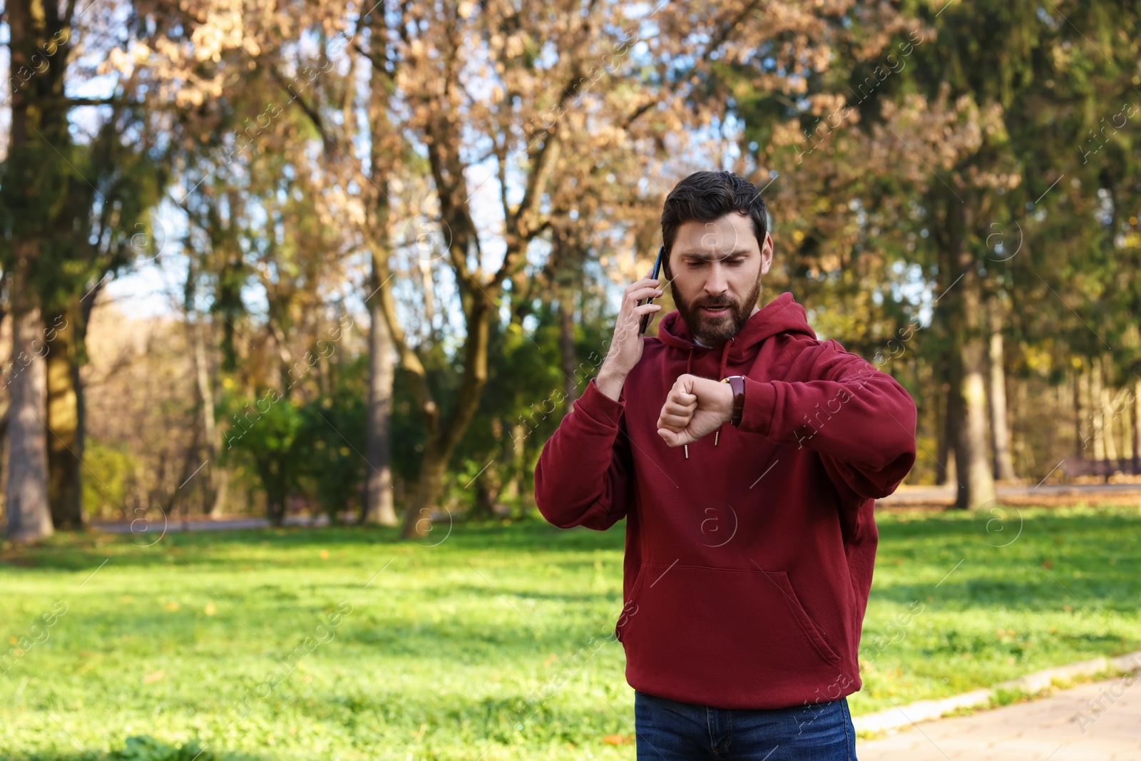 Photo of Emotional man talking by smartphone and checking time on watch in park, space for text. Being late concept