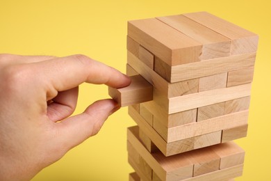 Photo of Playing Jenga. Man removing wooden block from tower on yellow background, closeup
