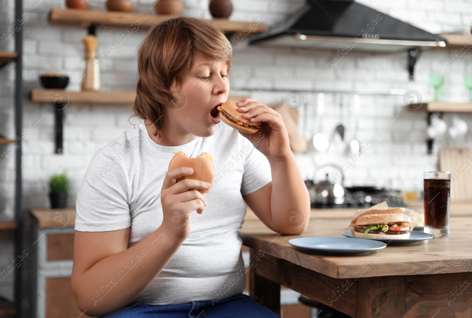 Photo of Overweight boy at table with fast food in kitchen