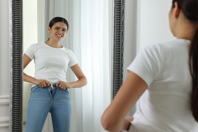 Photo of Young woman trying to put on tight jeans near mirror indoors