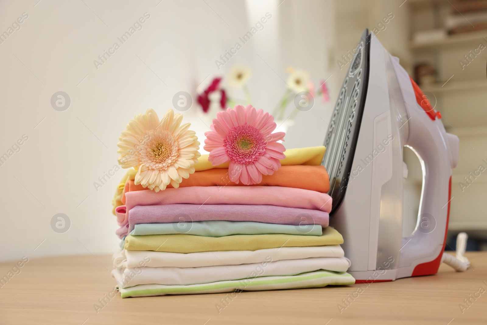 Photo of Stack of clean clothes, modern iron and flowers on wooden table