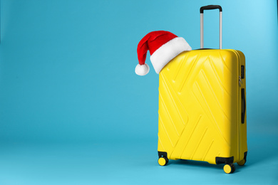 Image of Stylish suitcase with Santa Claus hat on blue background, space for text. Christmas vacation 