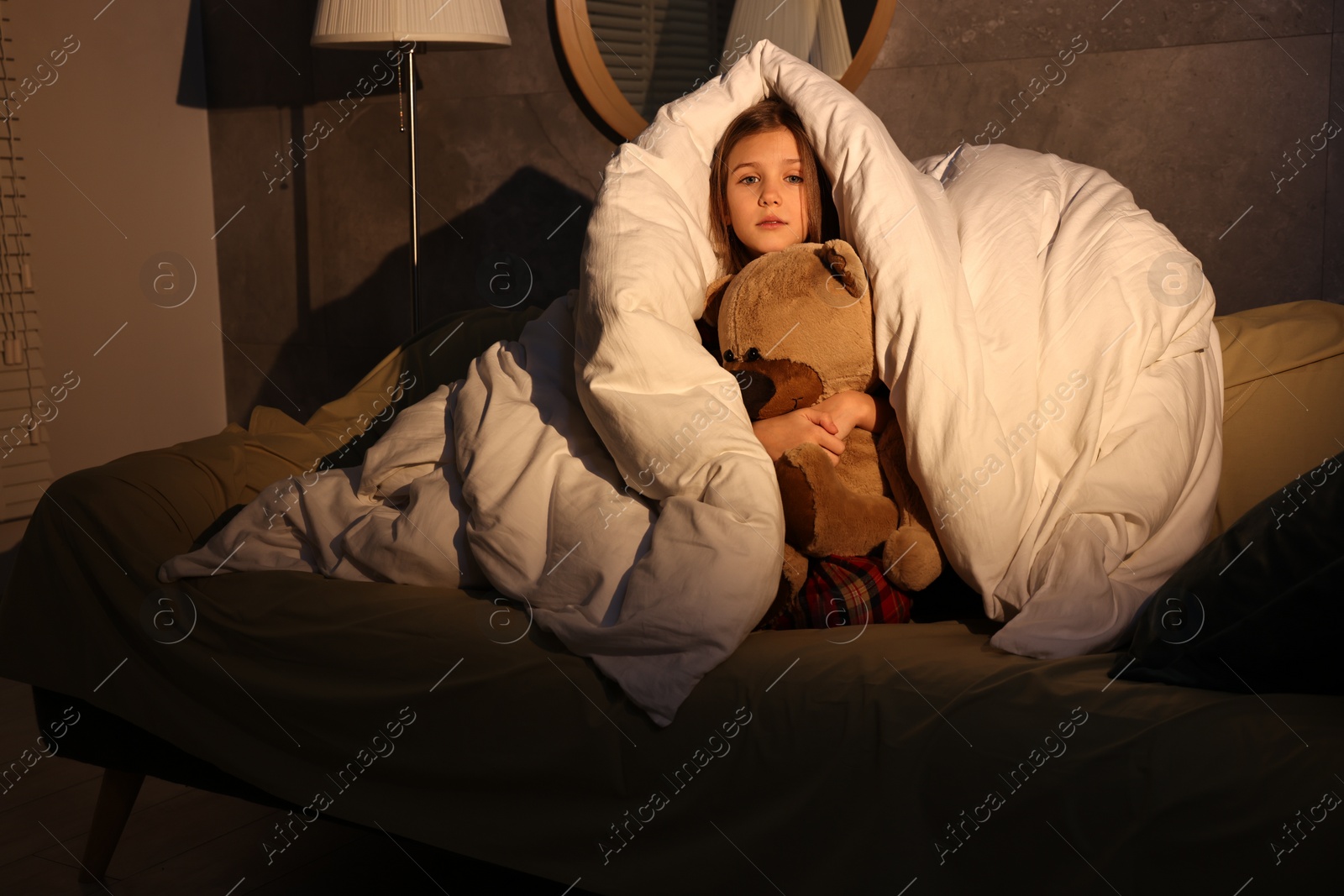 Photo of Scared little girl with toy hiding from monster in duvet on sofa at night, low angle view