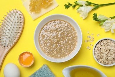 Photo of Homemade hair mask in bowl, ingredients and wooden brush on yellow background, flat lay