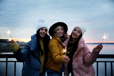 Women in warm clothes holding burning sparklers near river
