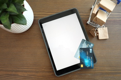 Photo of Internet shopping. Flat lay composition with small cart, credit cards and modern tablet on wooden table