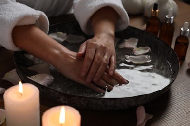 Photo of Woman soaking her hands in bowl of water and flower petals at wooden table, closeup. Spa treatment