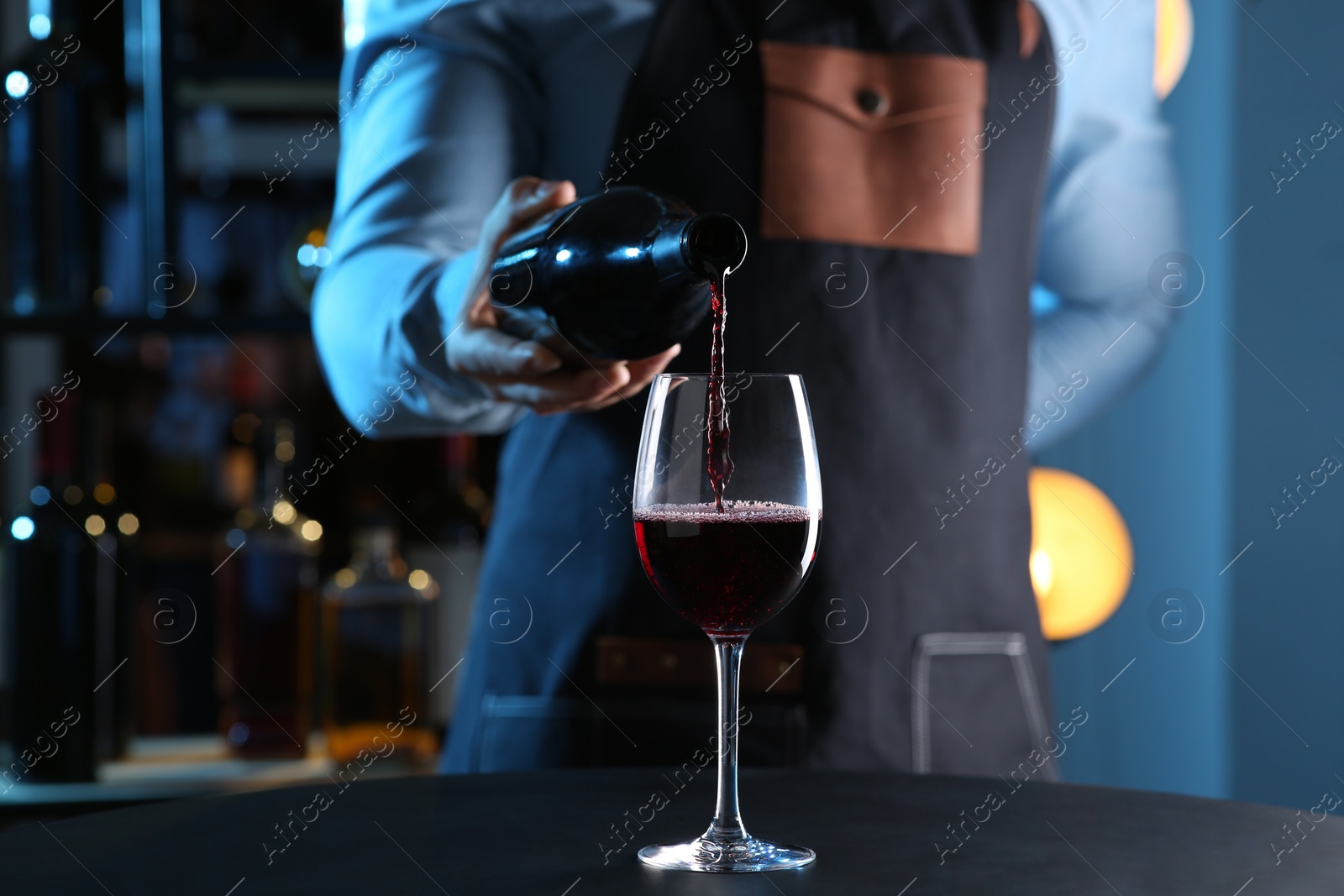 Photo of Bartender pouring red wine from bottle into glass at table indoors, closeup