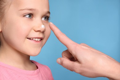 Photo of Mother applying ointment onto her daughter's nose on light blue background, closeup