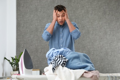 Photo of Emotional man near board with iron and pile of clothes at home