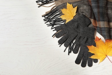 Stylish woolen gloves, scarf and dry leaves on white wooden table, flat lay. Space for text