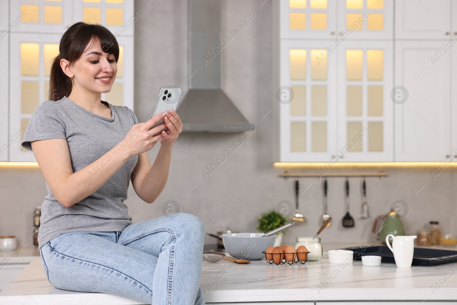 Photo of Happy young housewife using smartphone on white marble table in kitchen