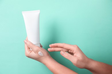 Woman with tube applying cosmetic cream onto her hand on turquoise background, closeup