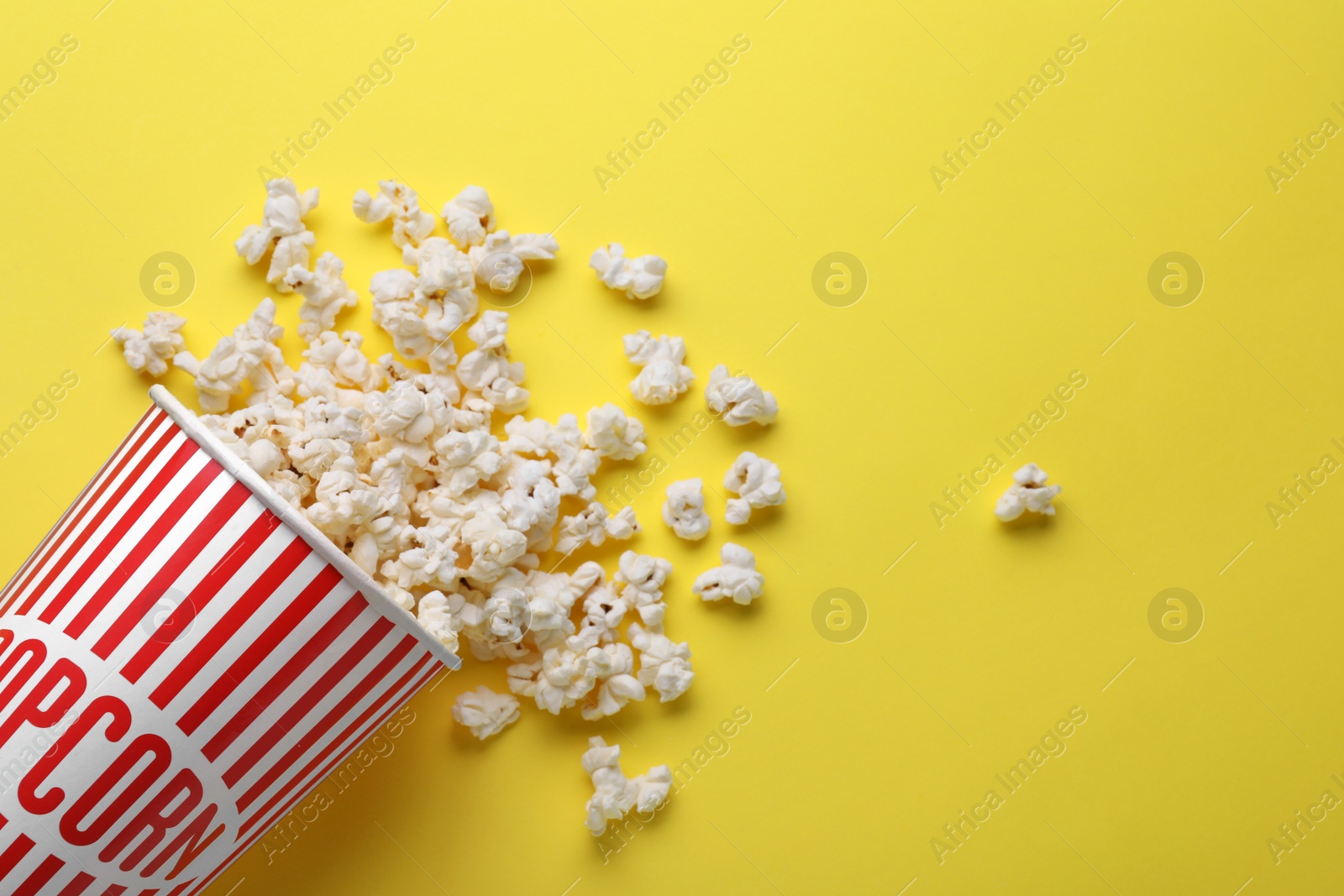 Photo of Overturned paper cup with delicious popcorn on yellow background, flat lay. Space for text