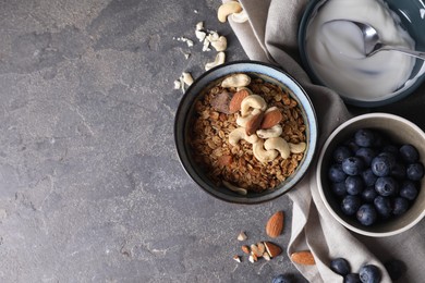 Photo of Tasty granola in bowl, blueberries, yogurt and spoon on gray textured table, flat lay. Space for text