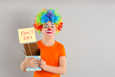 Photo of Preteen boy with clown makeup and wig holding APRIL FOOL'S DAY sign on light grey background, space for text