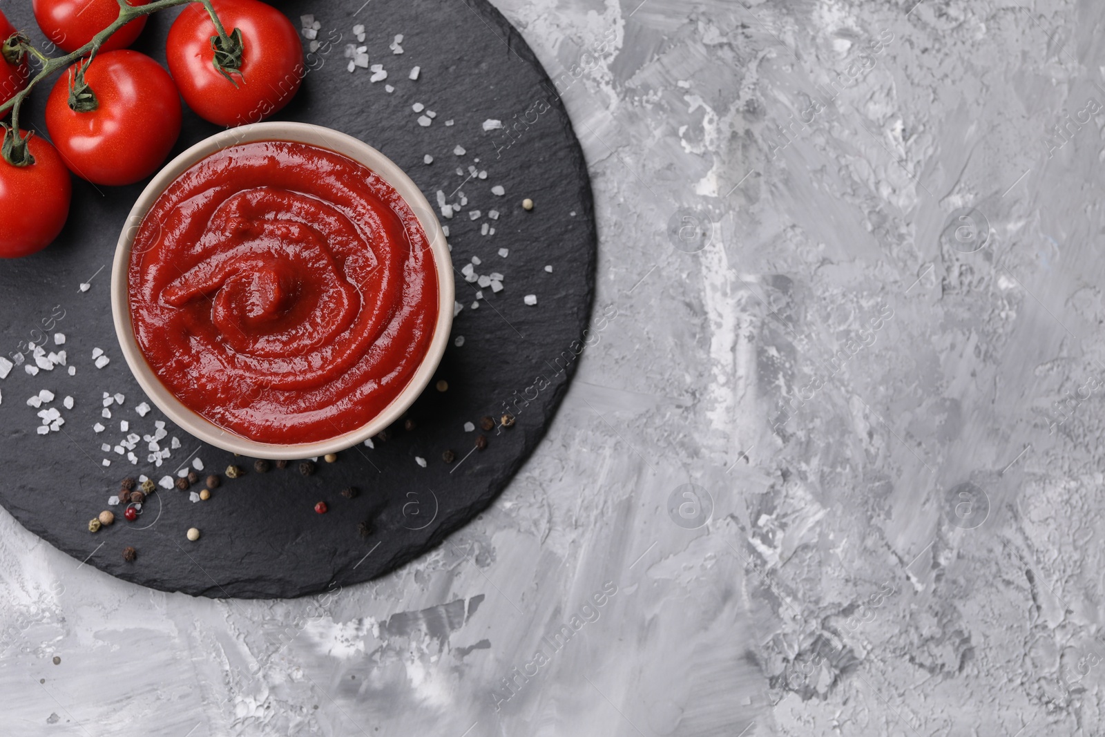 Photo of Organic ketchup in bowl, fresh tomatoes and spices on grey textured table, top view with space for text. Tomato sauce