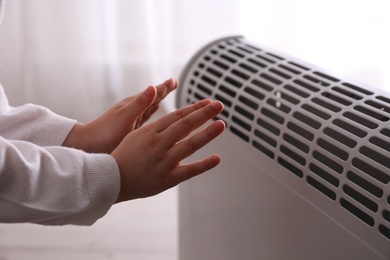 Photo of Child warming hands near electric heater at home, closeup