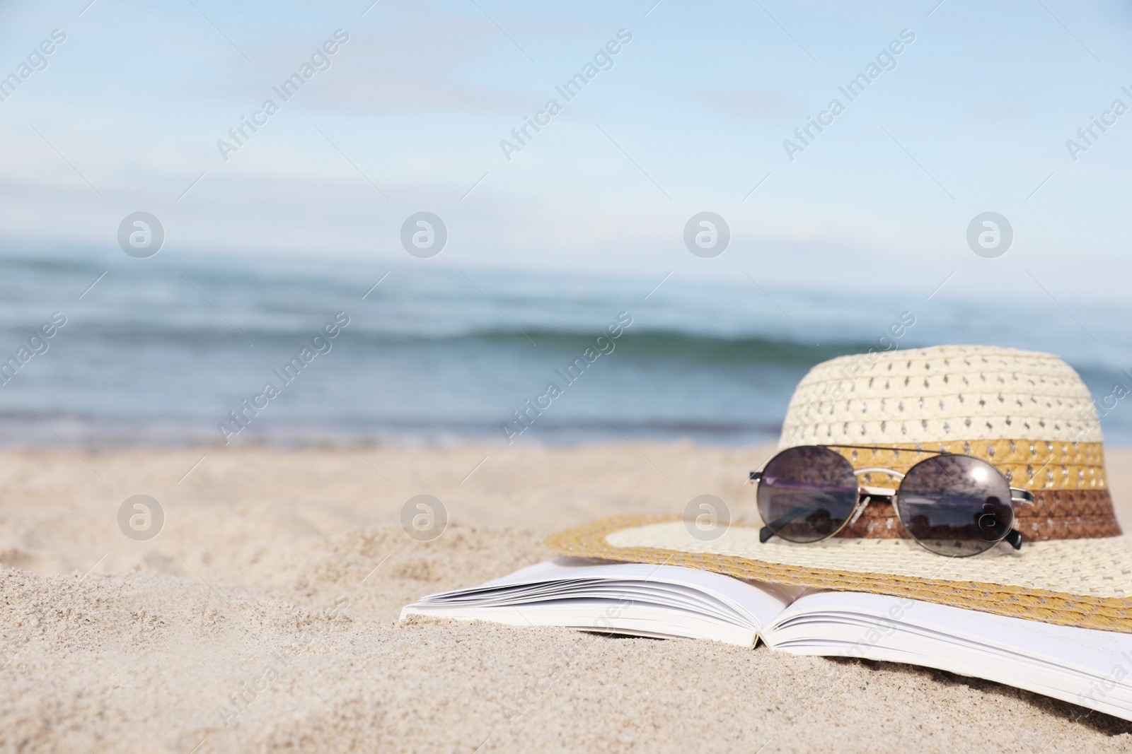 Photo of Open book, sunglasses and hat on sandy beach near sea, closeup. Space for text