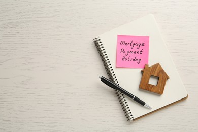 Photo of Paper note with words Mortgage Payment Holiday, notebook, pen and house model on white wooden table, top view. Space for text