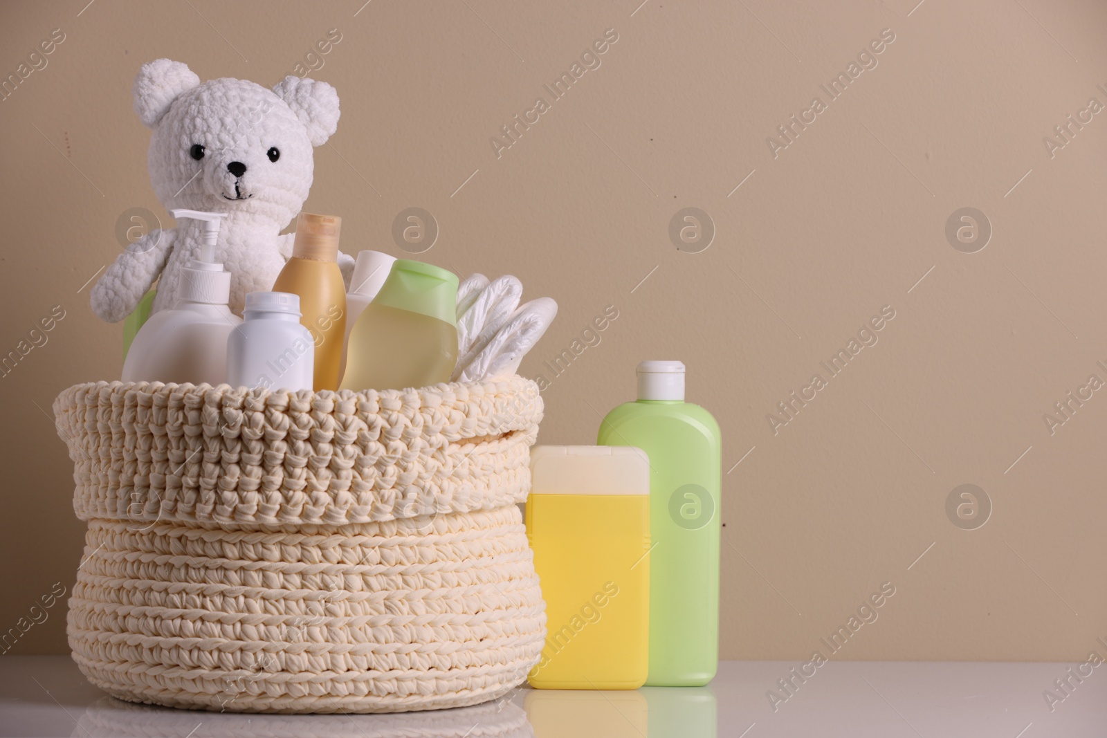 Photo of Knitted basket with baby cosmetic products and toy bear on white table against beige background. Space for text