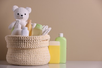 Photo of Knitted basket with baby cosmetic products and toy bear on white table against beige background. Space for text