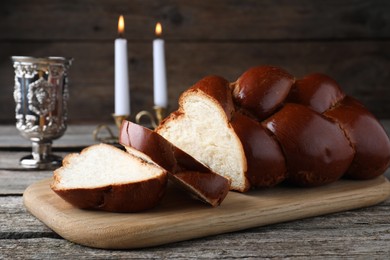 Photo of Cut homemade braided bread, goblet and candles on wooden table. Traditional Shabbat challah