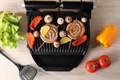 Photo of Electric grill with homemade sausages, vegetables and spatula on wooden table, flat lay