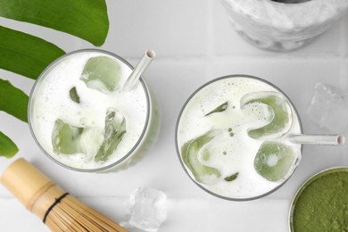 Photo of Glasses of tasty iced matcha latte, bamboo whisk and powder on white tiled table, flat lay