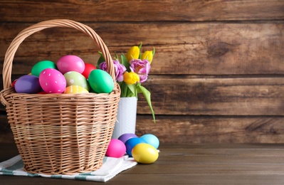 Photo of Colorful Easter eggs in wicker basket and tulips on wooden background. Space for text